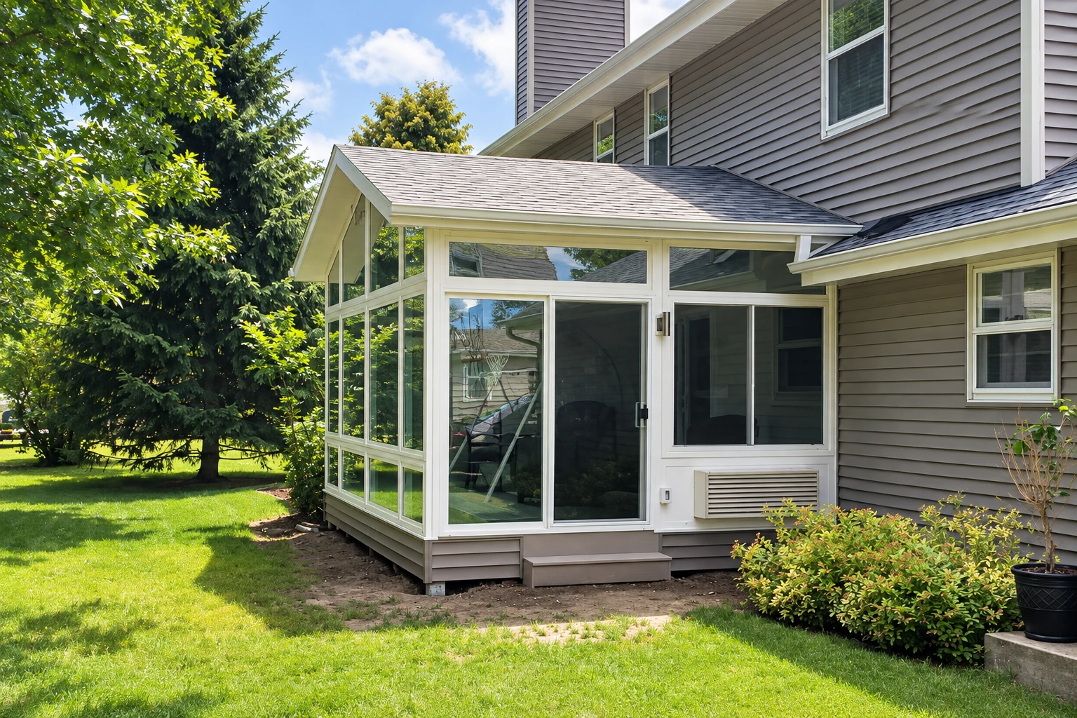 White-framed sunroom addition with a peaked roof, attached to a gray-sided house beside a green lawn.
