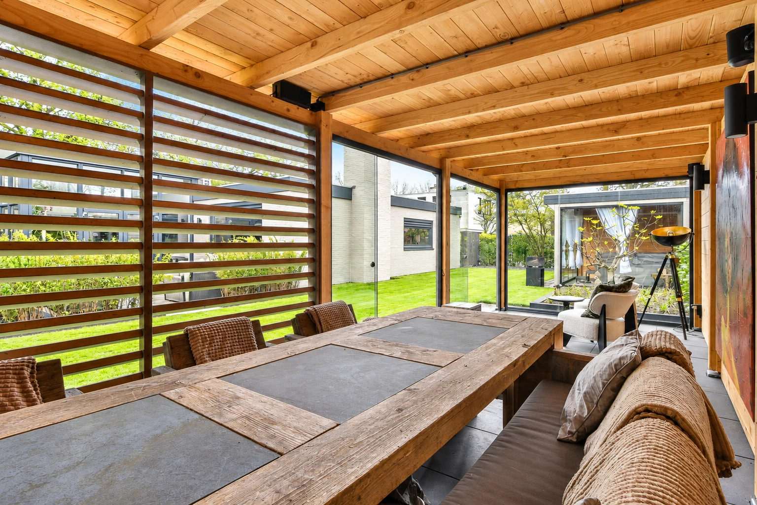 Enclosed patio room with a timber ceiling, slatted wood wall, long dining table and cushioned seating beside a lawn.