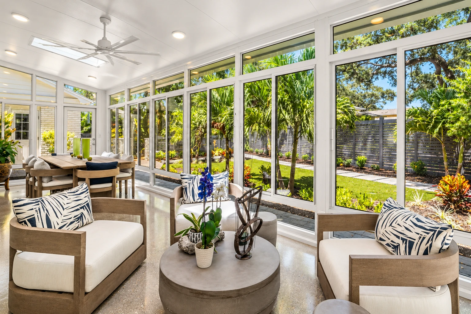 Four-season sunroom with white-framed windows, lounge chairs and a dining area overlooking a landscaped garden.